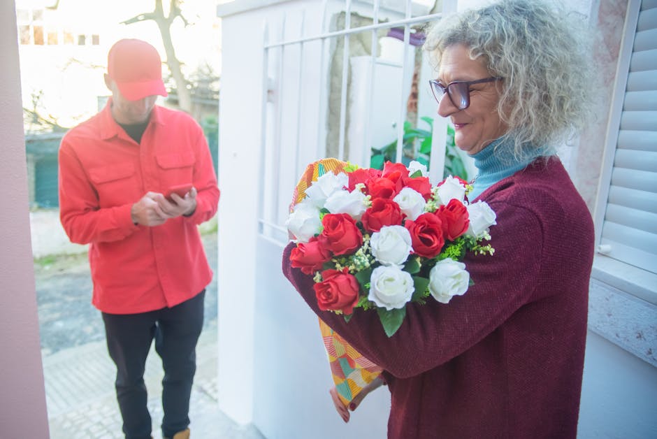A woman with curly gray hair, glasses, and a burgundy sweater is holding a vibrant bouquet of fresh red and white roses wrapped in colorful paper, standing outdoors near a white window with shutters a