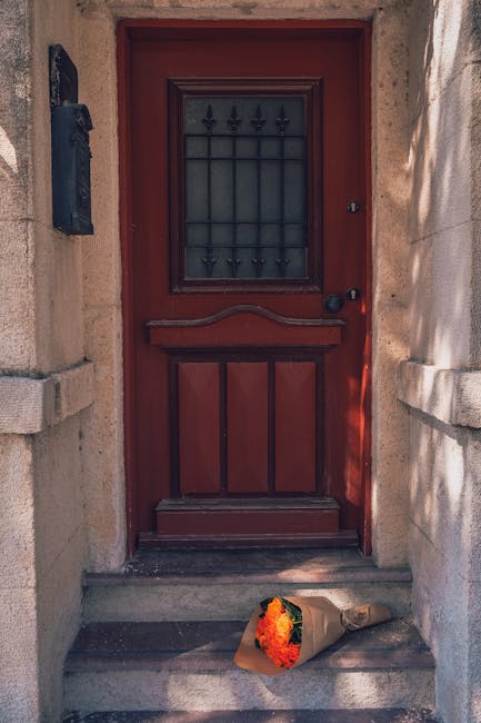 A bouquet of fresh flowers, featuring vibrant orange, yellow, and green hues, rests on the doorstep in front of a red wooden door with black iron accents and textured glass. The bouquet is wrapped in 