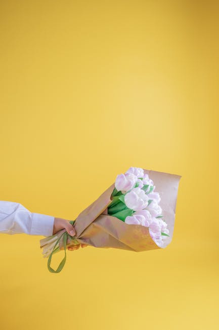 A person holding a bouquet of fresh light pink tulips wrapped in brown craft paper with a green ribbon, against a vibrant yellow background. The tulips have smooth, rounded petals and sturdy green ste