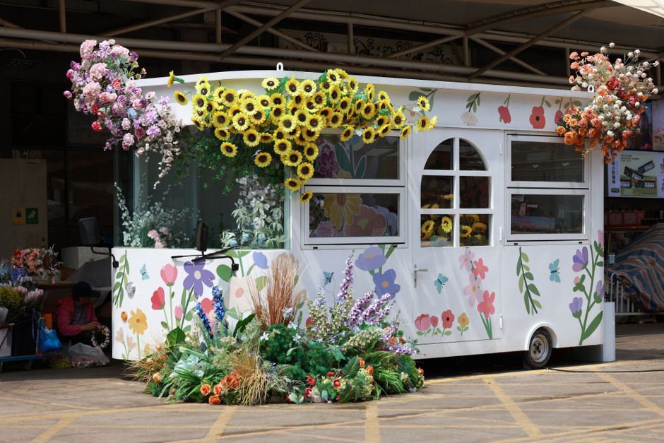 A white floral display kiosk decorated with cheerful, hand-painted floral illustrations in pink, purple, yellow, and green. The kiosk is topped with vibrant bouquets of fresh flowers, including large 