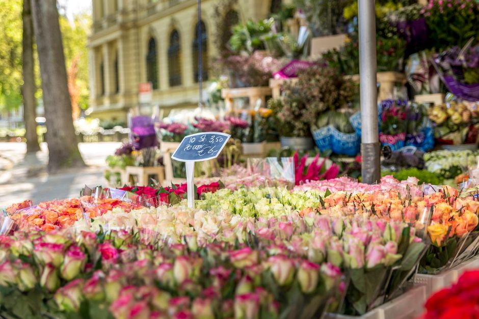 A vibrant outdoor flower stall featuring a variety of fresh, colorful flowers arranged in bouquets. The display includes roses in shades of pink, peach, and cream, wrapped in clear plastic, and arrang