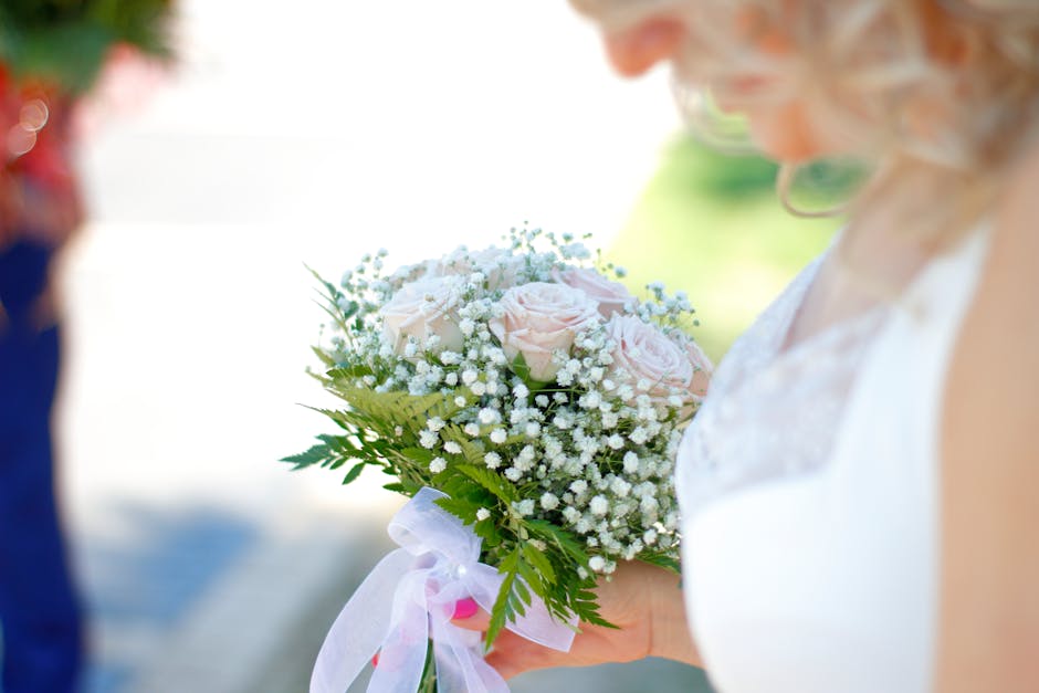 A close-up of a bridal hand holding a delicate bouquet consisting of pale pink roses and baby's breath, accented with lush green fern leaves. The bouquet is wrapped with a light pink satin ribbon, add