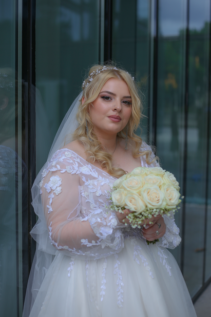 A bride with fair skin and long, wavy blonde hair styled with loose curls, wearing a delicate tiara and a sheer veil, holds a bouquet of ivory roses accented with small white flowers and greenery. She