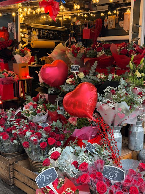 An indoor floral display featuring a variety of red and white flowers, including roses and carnations, arranged in bouquets wrapped in pink, white, and red tissue and paper. Large red heart-shaped bal